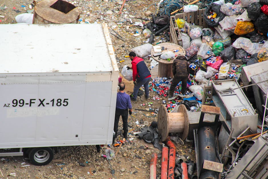 A large outdoor collection of overflowing rubbish bins and scattered waste materials situated on a paved area in front of a building with commercial storefronts. The scene includes various types of garbage bags, some black and others transparent, along with cardboard boxes, paper and plastic packaging, and miscellaneous household waste spilling onto the ground. A prominent grey bin labeled for mixed paper and cardboard is partially open, with magazines, cardboard, and paper protruding from the top. Next to it are black refuse bags and a bright red bin, all filled with waste. A small silver car is parked behind a metal railing on the left side of the image, and a tree is visible in the foreground, partially obstructing the view of the bins. The background features a multi-story building with a metal scaffold and blue safety netting, alongside shopfronts with signs indicating food and grocery outlets, suggesting an urban area where private waste collection services like Rubbish Collection Camden may be involved in handling excess rubbish outside regular collection times.