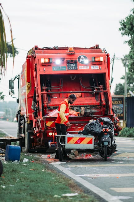 A worker dressed in a high-visibility orange and yellow vest is engaged in rubbish collection at the rear of a large red waste disposal truck parked alongside a roadside. The truck, with its rear loading mechanism visible, is positioned on the edge of the pavement, adjacent to a green grassy verge and a curb. The worker appears to be handling black plastic waste bags, which are placed on a small metal platform attached to the truck. Nearby, tools and equipment, including a small wheeled bin or container, are visible on the ground. The background shows some trees, utility poles, and a blue building with signage, indicating an urban or suburban environment. The scene is illuminated by natural daylight, providing clear visibility of textures such as the rough metal surface of the truck, the plastic bags, and the worker's fluorescent safety gear. This image exemplifies independent waste collection services, relating to non-local authority rubbish removal, often employed by private waste management providers like Rubbish Collection Camden for efficient on-site clearance of waste materials.