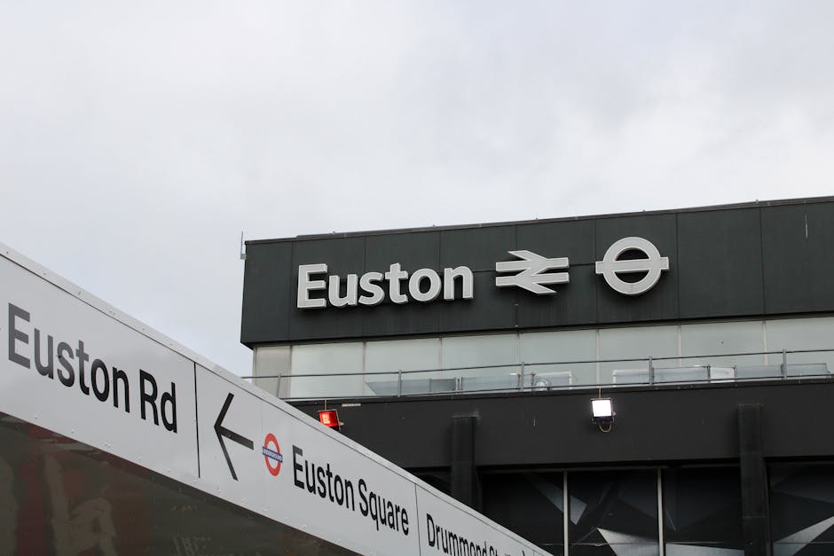 A daytime exterior view of the Euston railway station building, featuring the station name 'Euston' in large white letters on a dark grey facade. To the right of the station name, there are two white symbols: a double arrow indicating railway travel and a circle with an arrow pointing to the right. Below, a white sign with black text displays directions for Euston Road and Euston Square, with an arrow pointing left for Euston Road. The sky above is overcast with grey clouds, creating diffuse lighting across the scene. The surrounding environment suggests an urban setting, likely near a driveway or pavement area in front of the station, with clean and modern architectural elements. The signage and symbols serve as clear indicators for commuters and visitors, providing essential navigation information relevant to public transport and city travel, aligned with general themes of public transportation signage and access points related to rubbish and waste management services in the area.