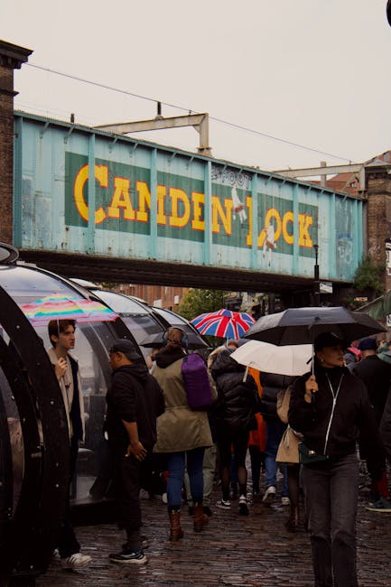 The image depicts a busy outdoor scene beneath a large blue railway bridge with the words 'CANDEN LOCK' painted in bold yellow letters. In the foreground, numerous pedestrians are gathered on a cobbled street, many holding umbrellas in various colours, including a prominent Union Jack design, indicating rainy weather. The crowd includes people wearing dark and neutral tones, with some engaged in conversation or walking past each other. To the left, a modern tram with a sleek, curved design and large windows is visible, reflecting the surroundings. The environment suggests an urban setting, likely near a transit hub or marketplace. The bridge above, made of weathered steel with visible rivets and supporting beams, spans across the scene, adding an industrial backdrop consistent with a railway or freight area. The scene's lighting is diffuse due to overcast skies, emphasizing the textures of the cobblestones, umbrellas, and the bridge structure. This setting aligns with local rubbish removal services managing waste in busy, high-traffic areas, possibly for event or street cleaning operations associated with Camden's public spaces.