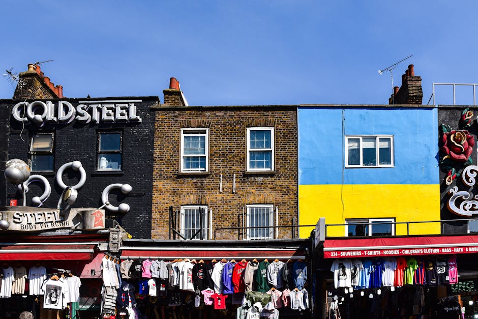 The image depicts a row of three adjacent buildings with contrasting exteriors. The building on the left has a dark black brick facade, featuring a large sign reading 'COLD STEEL' in white, three-dimensional letters. Below this sign, a smaller sign advertises a body piercing business, with some silver decorative elements and a red-and-white awning displaying various merchandise. The middle building has a traditional brown brick exterior with a slightly weathered appearance, featuring two windows on the upper floor, each with white frames and a grid pattern, and barred windows on the lower floor. To the right, a building is painted with a bright, two-tone color scheme: the upper half in sky blue and the lower half in vibrant yellow, divided by a black horizontal line. This building has two windows, one on each floor, with white frames. On the far right side, part of a mural is visible, with large, stylized roses and swirling patterns. Below the buildings, a street stall displays an assortment of clothing, including T-shirts, hoodies, and children's clothes, hanging on racks and laid out on tables, with a red canopy advertising souvenirs and various accessories. The scene is set against a clear blue sky, suggesting a bright, sunny day, and indicates a lively urban area that may be involved in independent or alternative waste disposal activities, with visible signs of rubbish collection services.